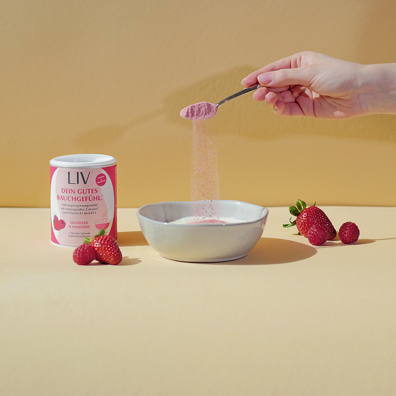 Hand gießt rosa Pulver aus einem Löffel in eine Schale mit Joghurt. Neben der Schale liegt die Verpackung der Darmkur und Erdbeeren und Himbeeren.