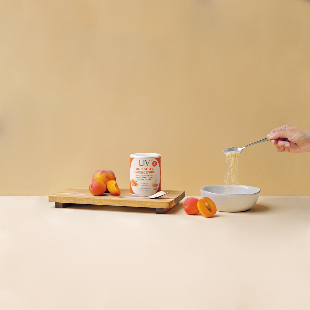Hand gießt gelbes Pulver  von einem Löffel in eine Schale mit Joghurt. Neben der Schale liegt die Verpackung der Darmkur und Pfirsiche und Aprikosen.